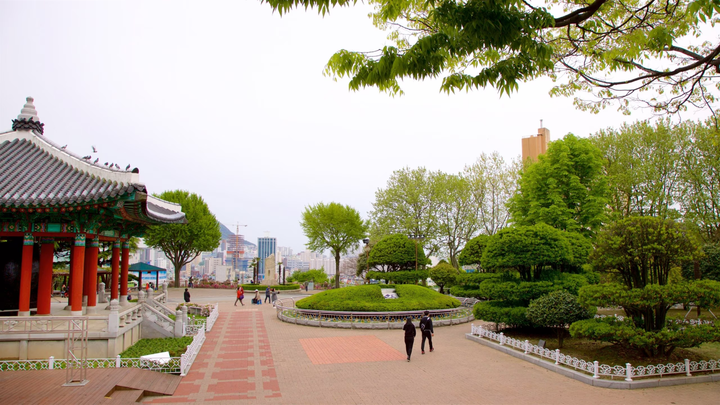 Região metropolitana, mostra construções com arquitetura oriental, prédios ao fundo, algumas árvores em uma praça publica com pessoas caminhando.